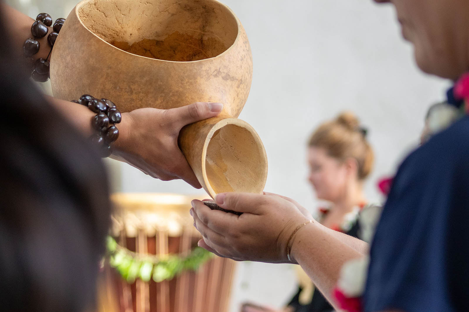 A close-up shows hands pouring liquid from a carved wooden vessel into another person’s cupped hands. Both wear leis and beaded bracelets, with blurred people and a drum adorned with leaves in the background. This header image is used on the "Our Leadership" page on the Nakupuna Foundation website. The Nakupuna Foundation is a Native Hawaiian Owned non-profit.