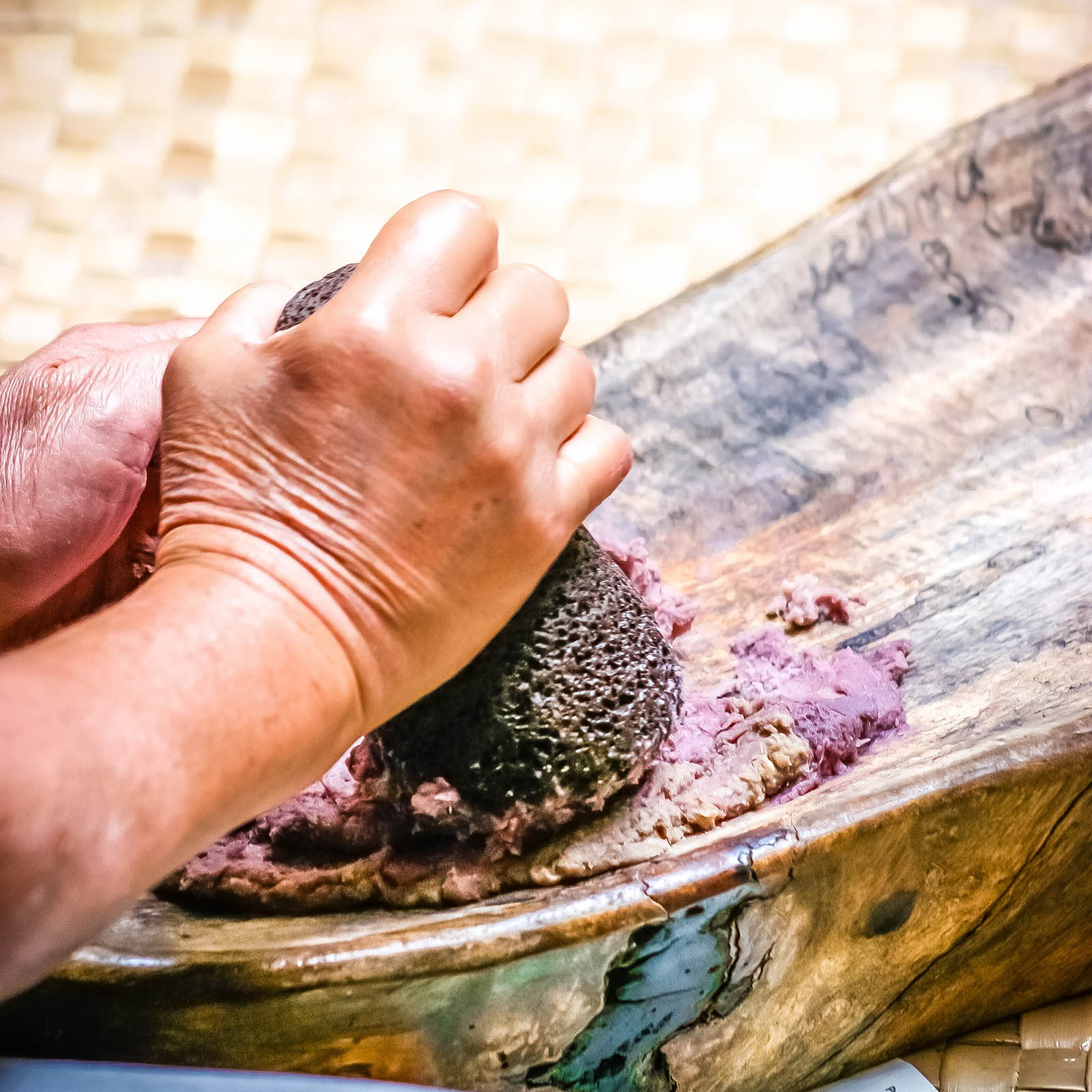 Close-up of hands using a stone tool to pound purple taro root on a wooden board, preparing poi. This header image is used on the "Our Purpose" page on the Nakupuna Foundation website. The Nakupuna Foundation is a Native Hawaiian Owned (NHO) non-profit.