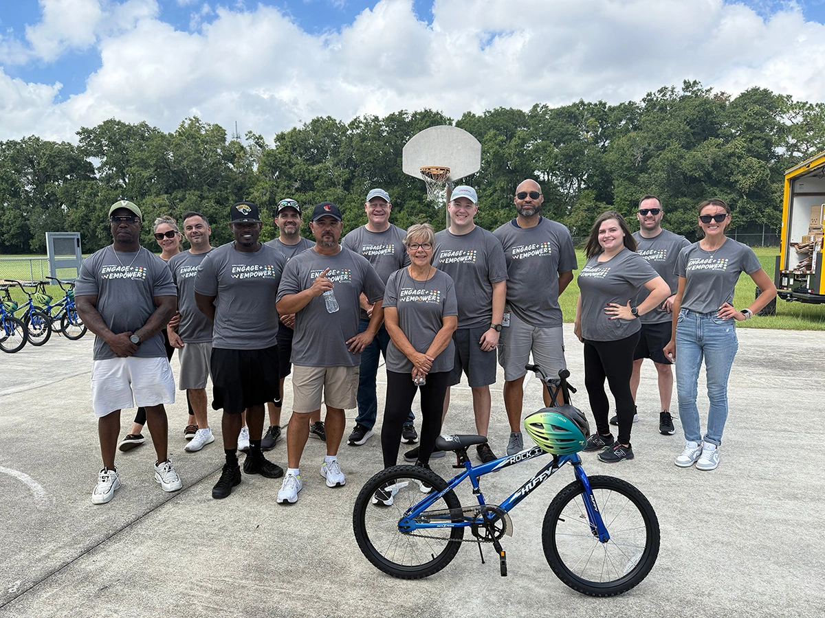 Group of volunteers with "engage empower" shirts stand with a blue kids' bicycle on a basketball court.