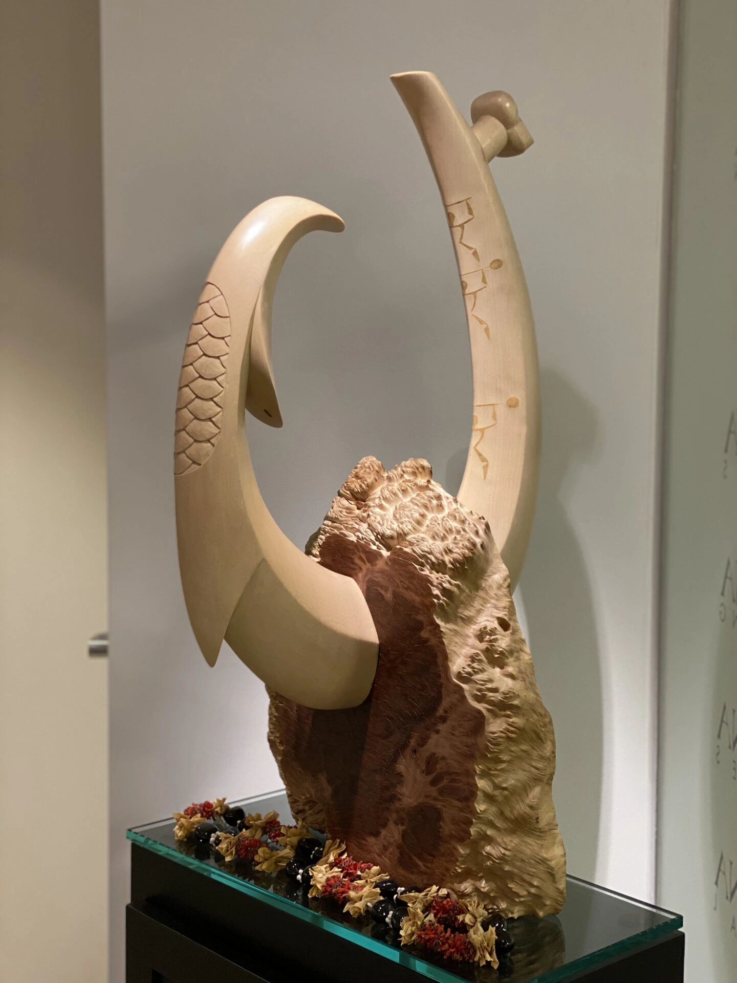 Carved wooden sculpture featuring a stylized fish hook emerging from a textured wood base, displayed on a glass table with floral accents, possibly representing nakupuna foundation's values.