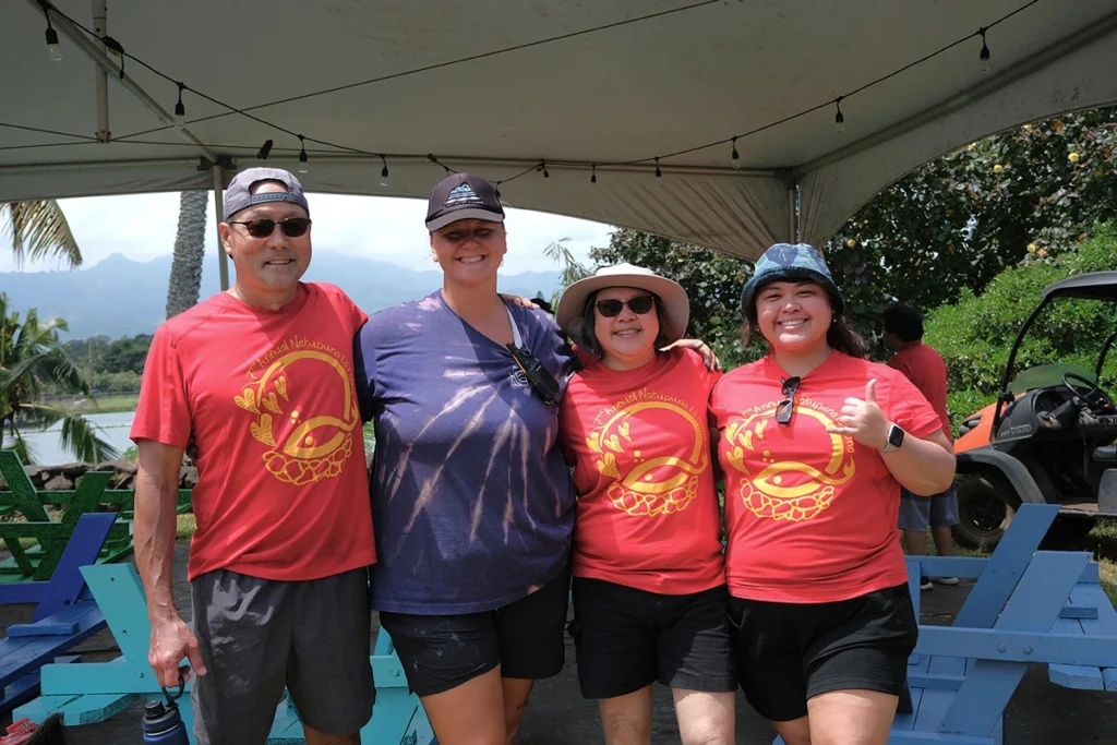 Our News 2 Nakupuna employees pose under a tent with string lights. Three wear red shirts with a gold design; one gives a thumbs up. Lush greenery, mountains, and a golf cart are visible in the background. This image is featured on the nakupuna foundation website.