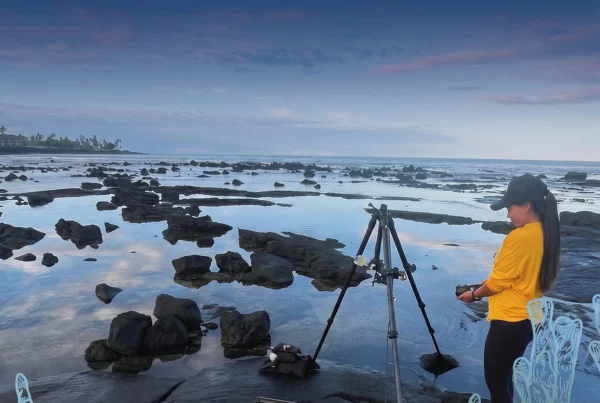 Two people are silhouetted against a twilight sky on a rocky Hawaiian shore, one working on a laptop near a tripod, the other holding a camera, hinting at community impact and advancement.