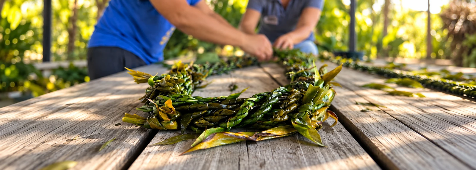 Two people's hands weave a vibrant green lei on a rustic wooden table outdoors, with lush foliage in the background.